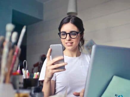 A woman wearing glasses sits at a desk with a laptop, looking at her smartphone. Art supplies are visible in the foreground as she manages multiple sites from her WordPress management for multiple sites dashboard. A woman wearing glasses sits at a desk with a laptop, looking at her smartphone. Art supplies are visible in the foreground as she manages multiple sites from her WordPress management for multiple sites dashboard.