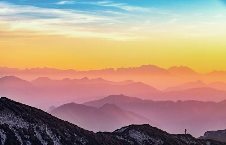 A silhouetted person stands on a mountain ridge overlooking layered mountain ranges at sunset, with a colorful sky above.