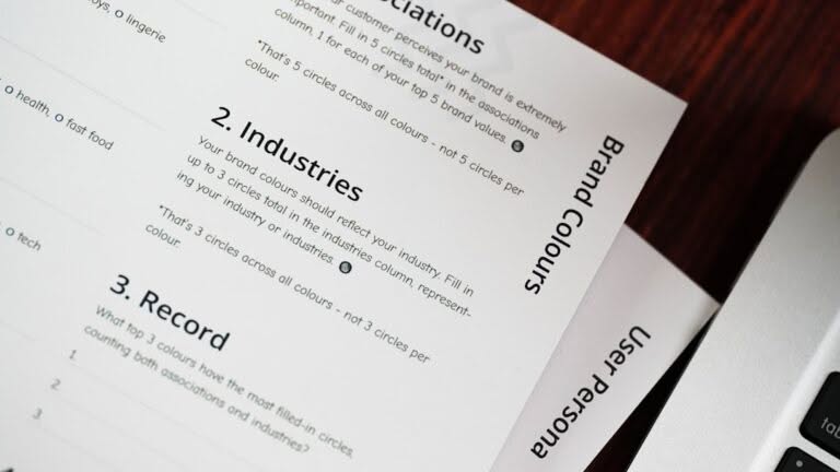 Close-up of printed documents showing sections titled "Industries," "Record," "Brand Colours," and "User Persona" next to part of a laptop keyboard.