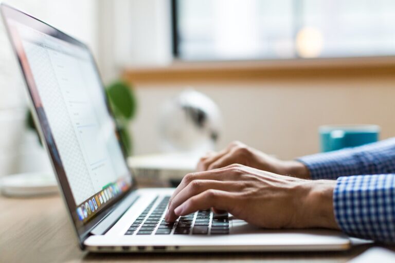 A person wearing a blue checkered shirt types on a laptop keyboard near a window with a blurred background.
