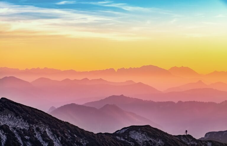 Colorful sunrise or sunset over layered mountain ridges with a silhouetted person standing on a rocky outcrop in the foreground.