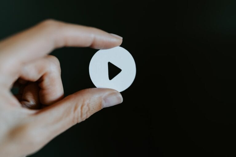 A hand holds a small white circular cutout with a black play button icon in the center against a dark background.