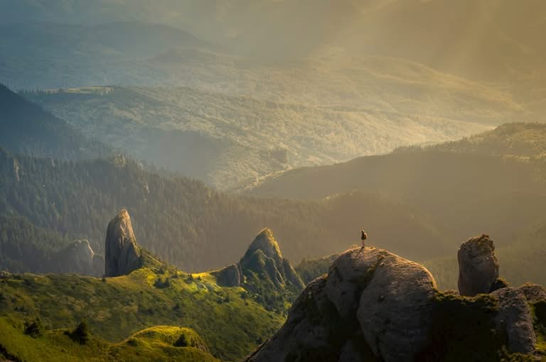 A person stands on a rocky peak overlooking rolling green hills and sunlit mountains covered in forest.